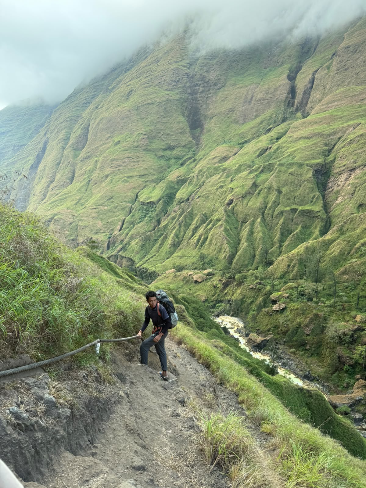 A hiker climbing the steep roped section of the Torean canyon trail, the green gorge wall rising behind