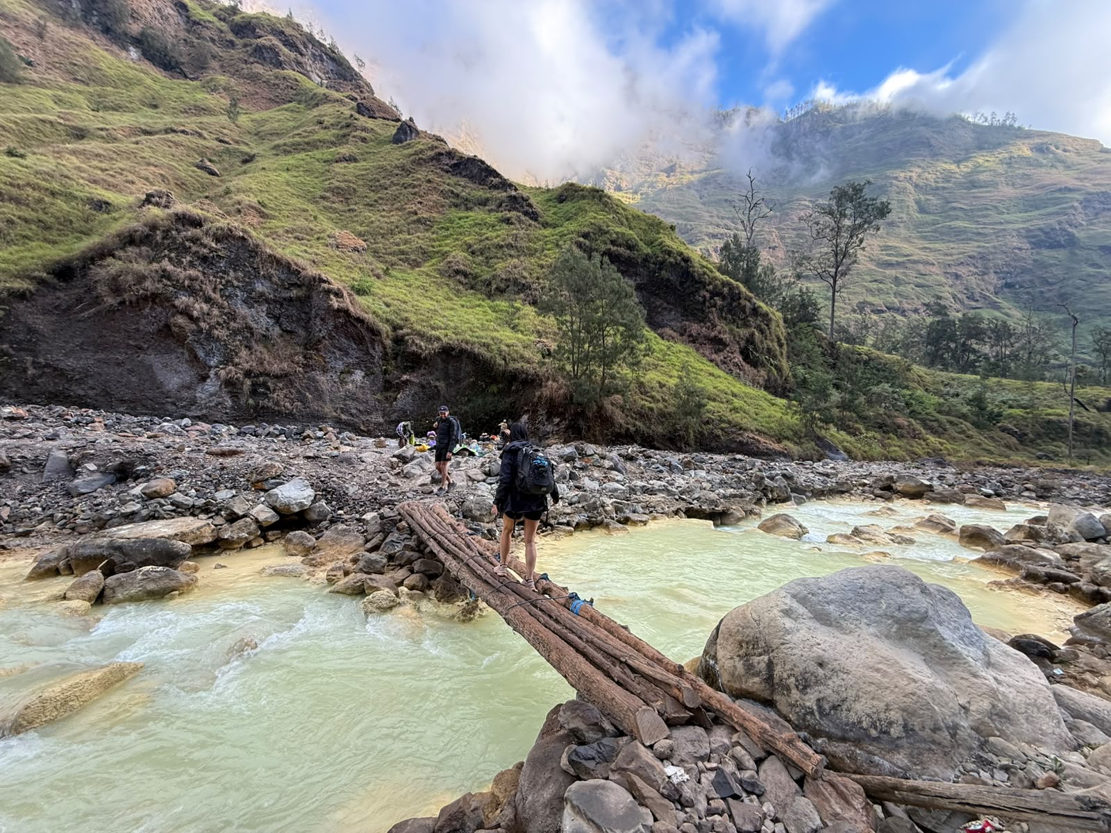 A trekker crossing the timber footbridge over the turquoise Torean river, the group resting on the far bank